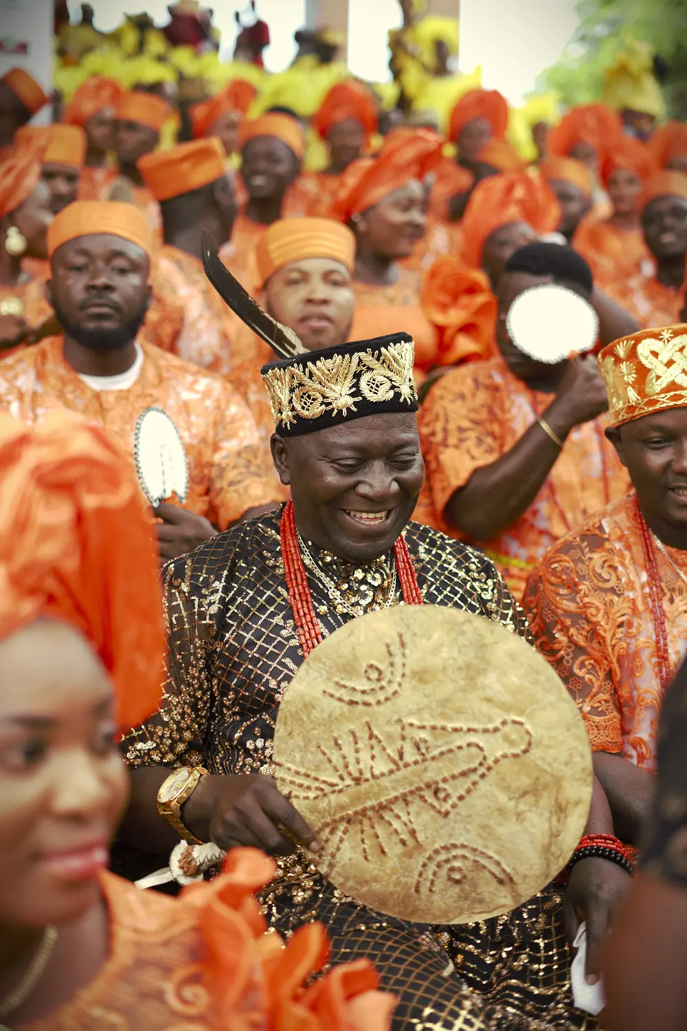 A smiling elder in black and gold with a leather fan, among dancers in orange